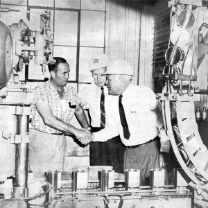 Two white men in shirt and tie and helmets greeting man in working clothes amid machinery indoors