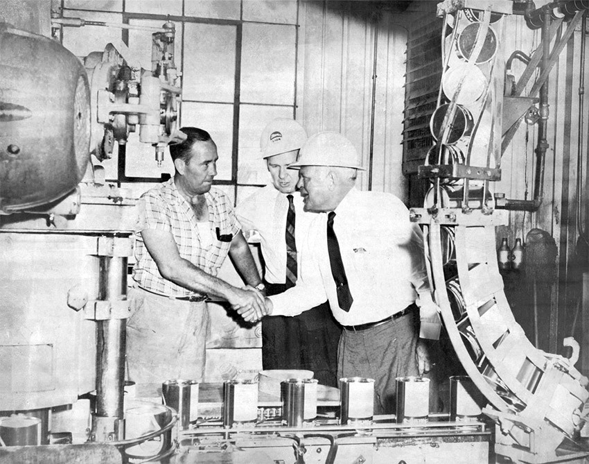 Two white men in shirt and tie and helmets greeting man in working clothes amid machinery indoors