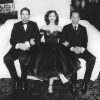 White woman in off-shoulder dress sitting on couch between two white men in suit and tie