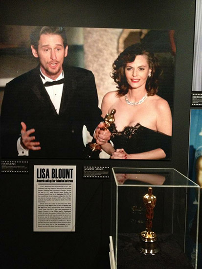 Gold statue in museum display case with photograph of white man in suit and white woman in off-shoulder dress above it