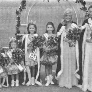 Group of young white girls in dresses robes and tiaras standing in line with arch behind them