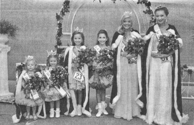 Group of young white girls in dresses robes and tiaras standing in line with arch behind them
