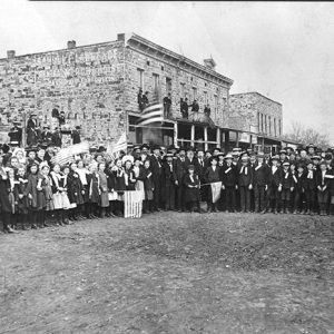 Crowd of white men women and children with some holding American flags on dirt road with multistory brick buildings behind them