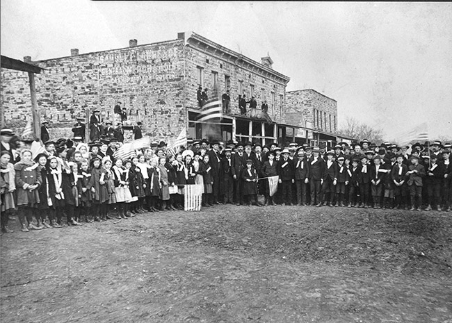 Crowd of white men women and children with some holding American flags on dirt road with multistory brick buildings behind them