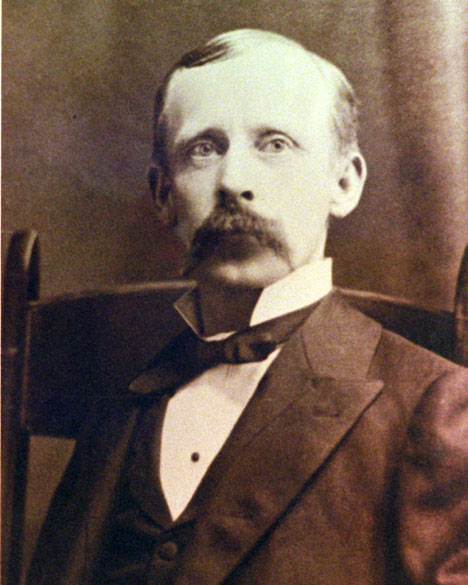 White man in suit with large mustache sitting in wooden chair