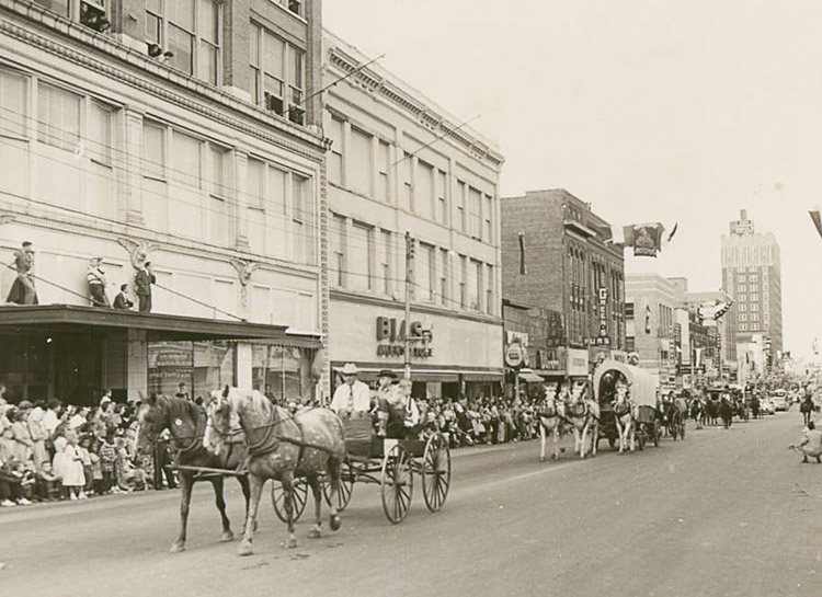 Horse-drawn wagons in parade on city street with crowds of people standing in front of multistory buildings