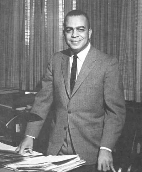 African-American man in suit and tie smiling at his desk