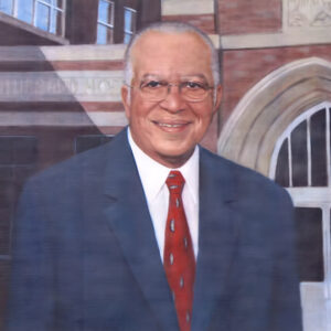 African-American man with glasses in suit and tie with multistory brick building behind him