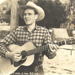 White man in western clothing and hat playing an acoustic guitar