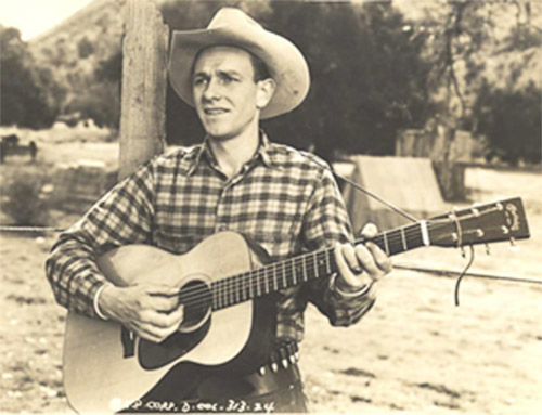 White man in western clothing and hat playing an acoustic guitar