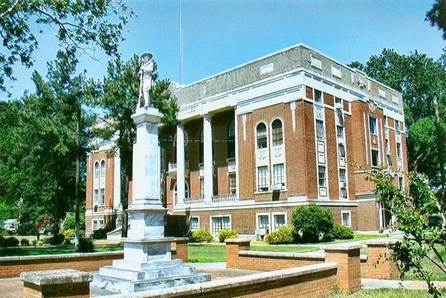 Monument on pedestal with statue on top in front of multistory brick building with white columns