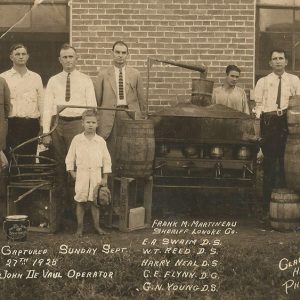 Group of white men and white boy posing with moonshine still outside brick building