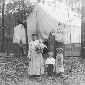 White woman holding baby alongside young boy and girl standing before a wooden structure covered by a tent