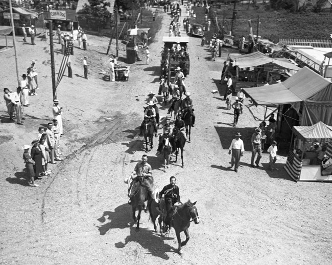 Parade of people on horses with spectators watching on a dirt road