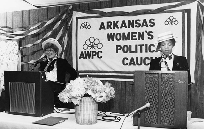 White woman and African-American woman speaking at lecterns on table with "Arkansas Women's Political Caucus" banner on the wall behind them