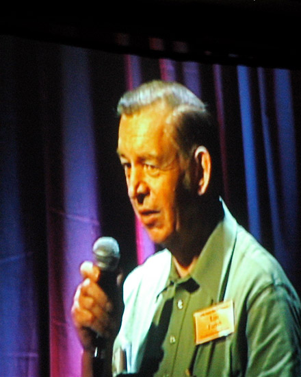 Old white man speaking into microphone in green shirt