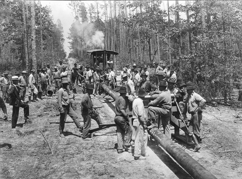 Group of men in woods beside large pipe with steam-powered vehicle in distance