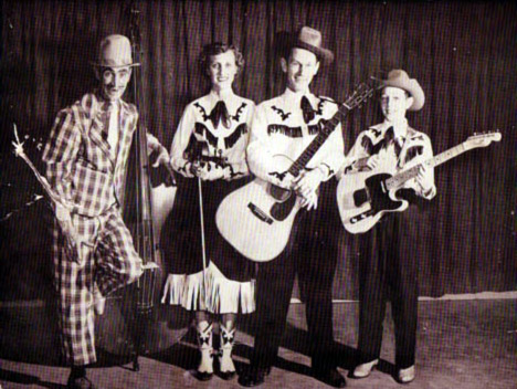White man and woman with boy in western clothing alongside white man in checkered suit, all standing on a stage and holding instruments