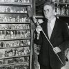 Older white man with glasses in suit standing before shelves filled with various items