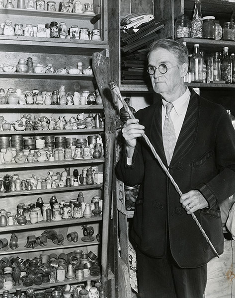 Older white man with glasses in suit standing before shelves filled with various items