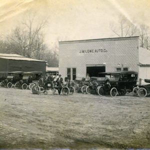 White men and cars outside "J. W. Lower Auto Company" building and "Lumber" building on dirt road