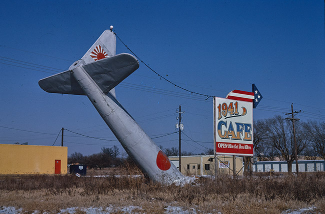 Tail of airplane with Japanese imperial flag painted on it sticking out of the ground next to "1941 Cafe" sign