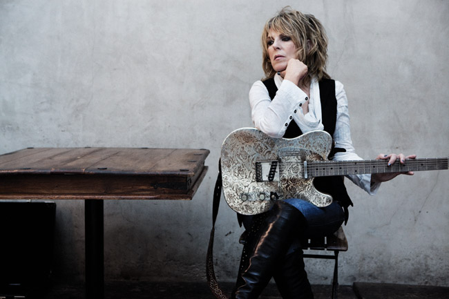 White woman with black and white outfit and heavy eye makeup sitting at a wooden table holding a silver Fender Telecaster guitar
