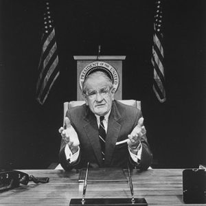Old white man with glasses in suit and tie talking at desk with lectern and flags behind him
