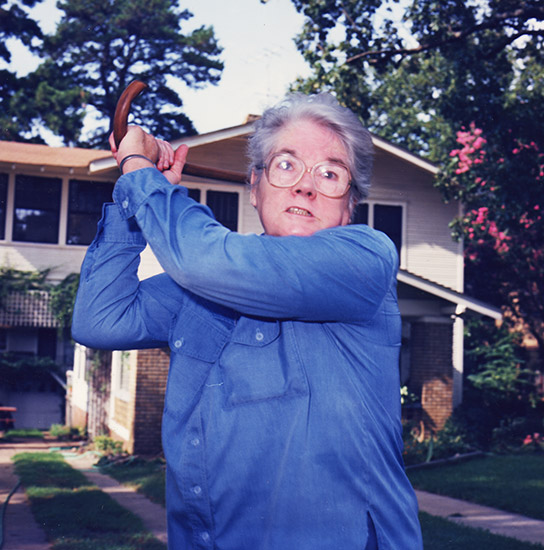 Older white woman brandishing her cane with two-story house behind her