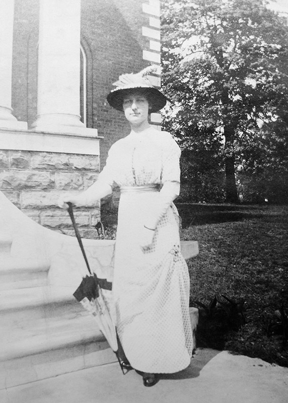 White woman in hat and dress leaning on closed parasol outside brick building