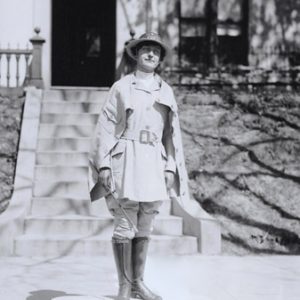 White woman wearing a uniform hat and boots standing with house and steps behind her