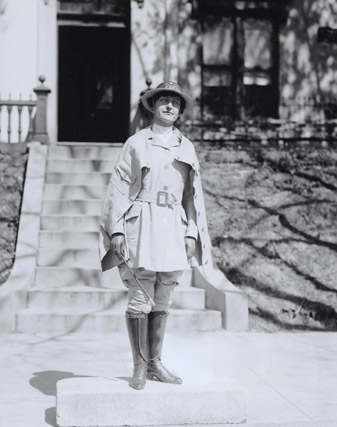 White woman wearing a uniform hat and boots standing with house and steps behind her