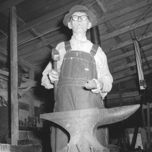 White man in overalls holding hammer behind anvil