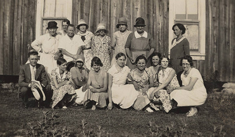 White man in suit and tie sitting with group of white women in dresses outside church building