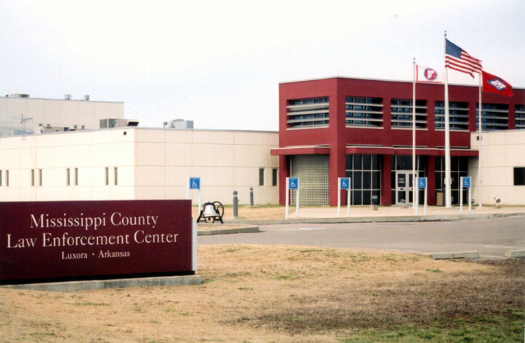 Concrete building with three flags, a bell and a sign "Mississippi County Law Enforcement Center Luxora Arkansas" in front