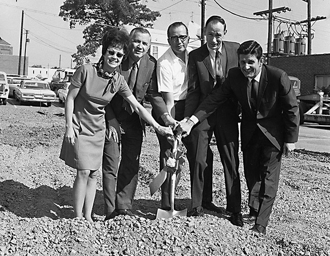 White men in suits with white woman in dress and sunglasses posing with hands on a shovel in dirt