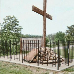 Large cross in pile of rocks with plaque surrounded by iron fence