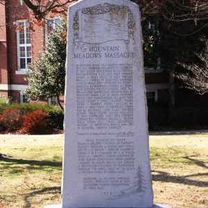 white stone monument with pointed top labeled "The Mountain Meadows Massacre" in front of multistory brick building