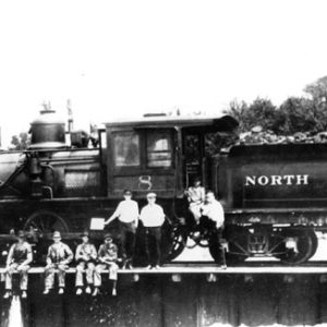 Steam locomotive with coal car saying "North Arkansas" on the side and white workers standing and sitting on the tracks