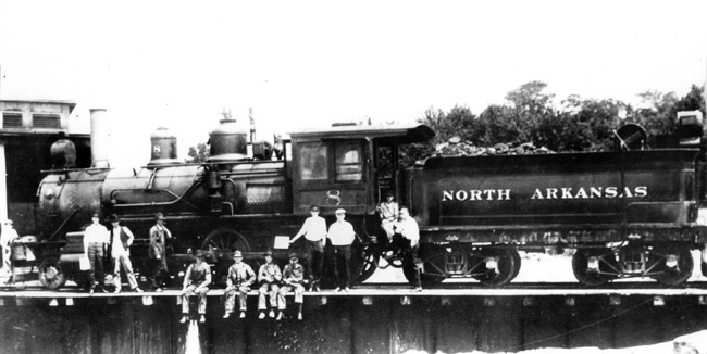 Steam locomotive with coal car saying "North Arkansas" on the side and white workers standing and sitting on the tracks