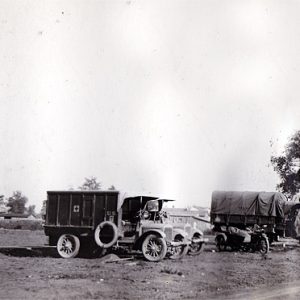 Military trucks on dirt with buildings and trees in the background