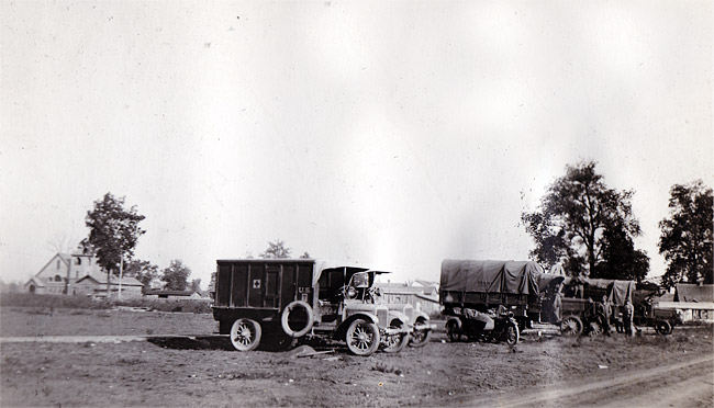 Military trucks on dirt with buildings and trees in the background