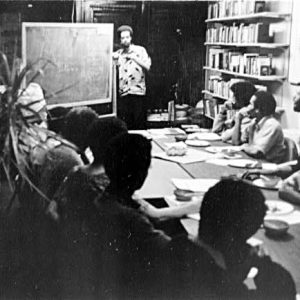 mostly African American group of men sit around table surrounded by filled bookshelves and looking at instructor standing next to chalk board