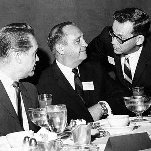 Three white men in suits talking at table with drinks and plates