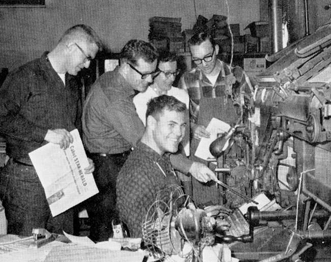 Group of white men with linotype machine, some holding issues of a newspaper