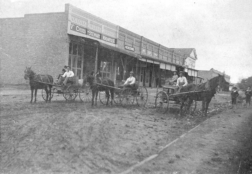 Men in three horse drawn buggies on dirt road with storefronts on left side