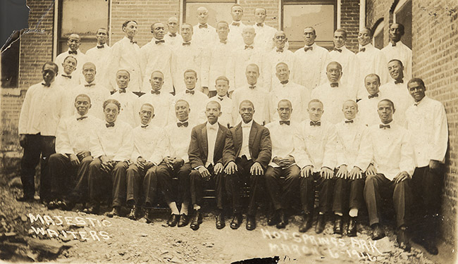Group of African-American men in matching uniforms with white jackets and bow ties and two in front with matching black suit jackets posing in front of brick building