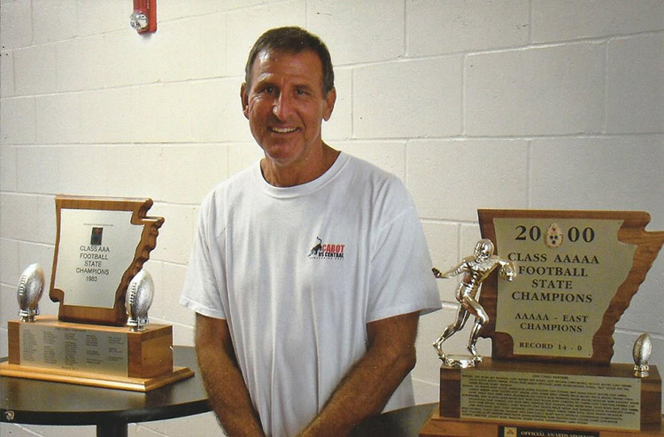 White man smiling in white shirt with Arkansas shaped trophy on table next to him