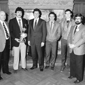 Group of white men in front of fireplace with one holding a trophy