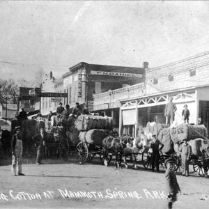 Men transporting cotton bales with horse-drawn wagons on town street with multistory storefronts